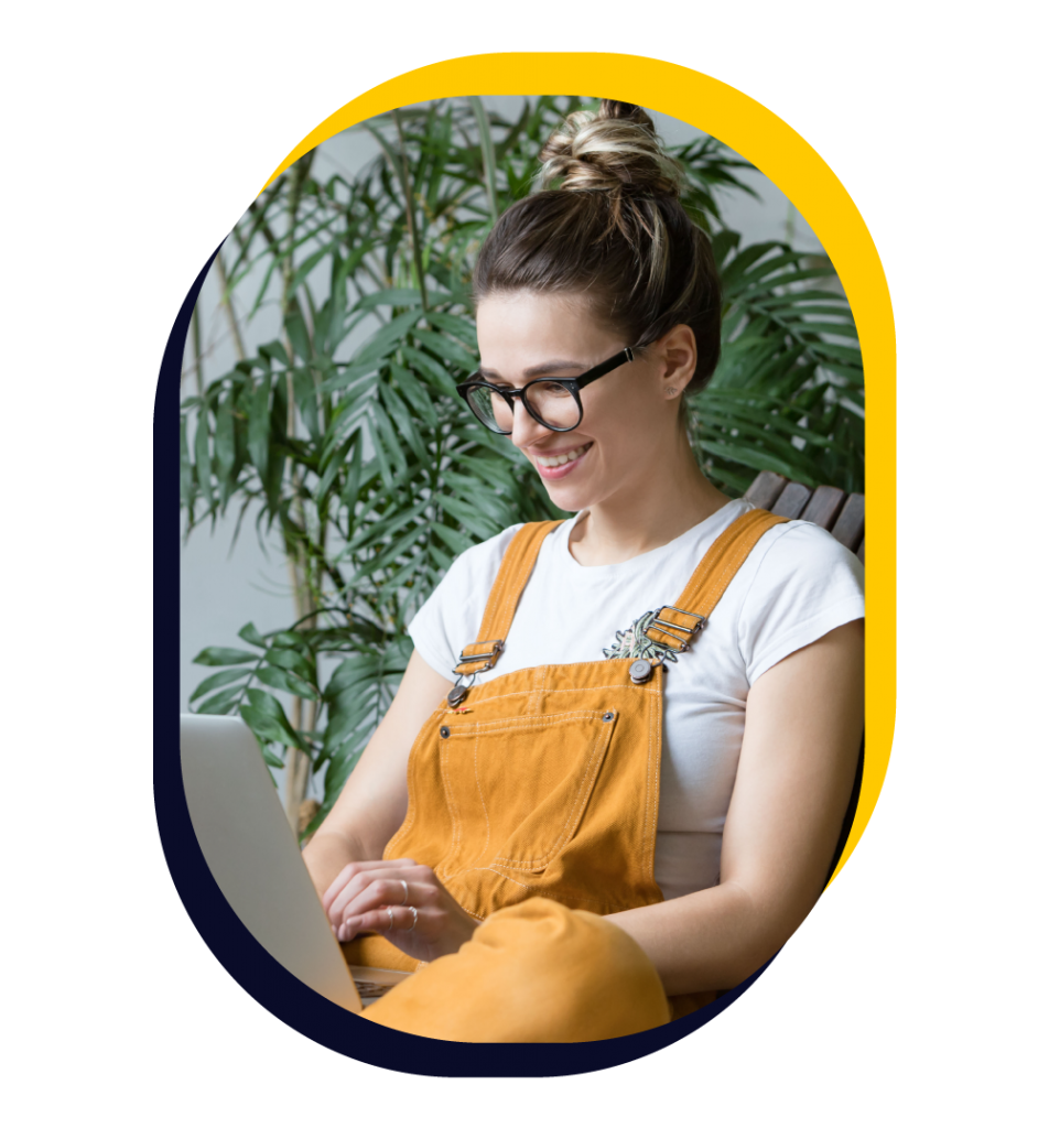 Woman in yellow overalls and a white t-shirt applying to a course on her laptop while sitting in front of a house plant