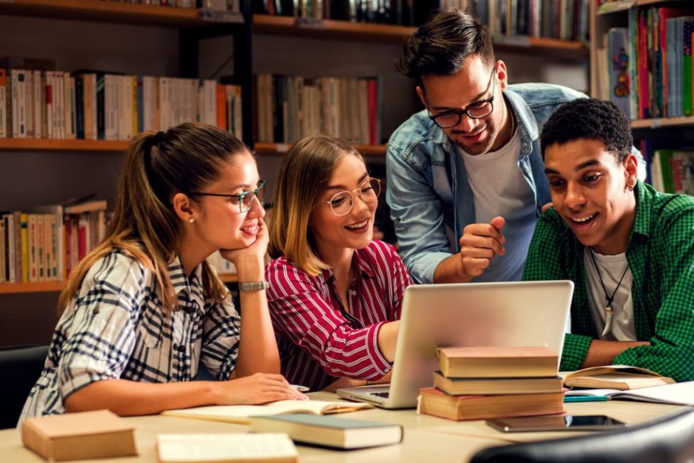 Four young students study in the school library, female student using laptop for researching online.